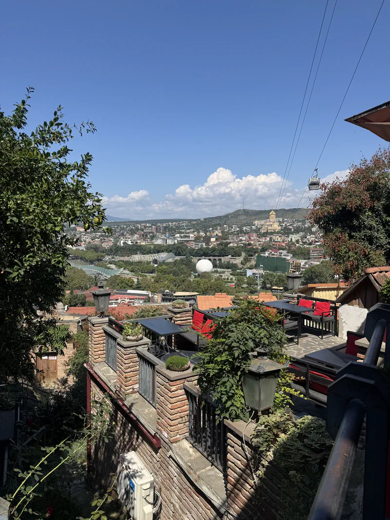 The terrace at 144 Stairs on a sunny day, with red cushions, plants and a wide view over Tbilisi and the cable-car line.